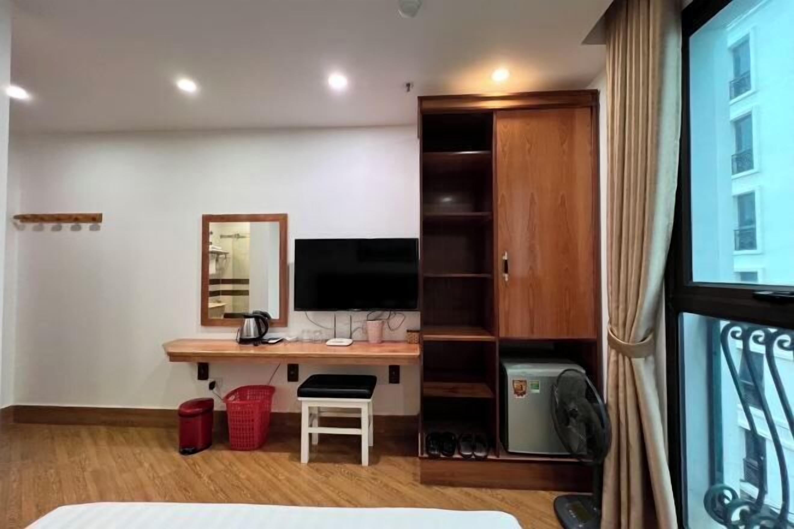 Una habitación de hotel con un escritorio de madera, televisor, armario, cama y ventana con cortinas.