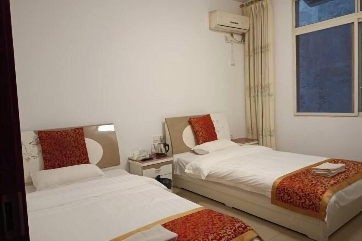 Ein Hotelzimmer mit zwei Einzelbetten, einem kleinen Nachttisch mit Lampe, einer Klimaanlage und einem Fenster mit Vorhängen.