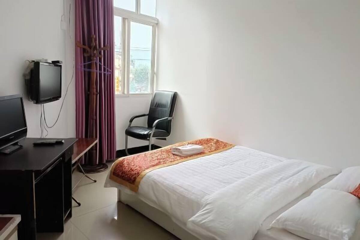 Ein Hotelzimmer mit einem Bett, einem Schreibtisch mit Fernseher, einem Sessel und einem Fenster mit Vorhängen.