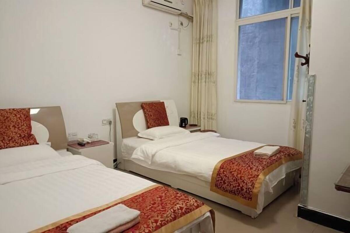 Ein Hotelzimmer mit zwei Betten, jeweils mit einer rot-weißen Tagesdecke, einem Nachttisch mit Lampe und einem Fenster mit Vorhängen.