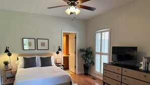Un dormitorio con cama, mesitas de noche, ventilador de techo, televisión, cómoda y una puerta que da a otra habitación.