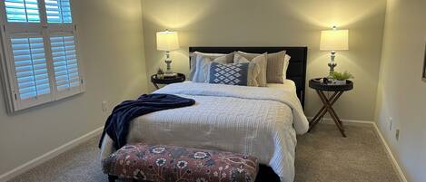 Un dormitorio con cama, dos mesitas de noche con lámparas, una ventana con persianas y suelo alfombrado.