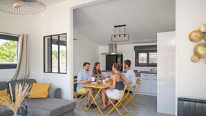 A modern living room with a grey sofa, yellow cushions, and a glass vase with dried plants. A dining table with yellow chairs is set for four people. The room has large windows and a view of greenery outside. - Comfortable 3-Bedroom Vacation Rental: NUIKI Lodges (Montségur-sur-Lauzon)