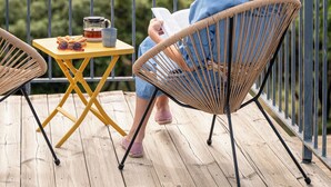Une personne assise sur une chaise en osier, sur une terrasse en bois, devant une table, et qui lit un livre.