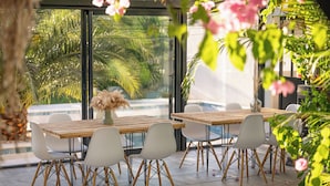 Een terras met een houten tafel en witte stoelen, omgeven door grote ramen met uitzicht op palmbomen en een zwembad.