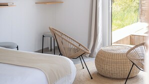 Een moderne slaapkamer met een bed, twee rieten stoelen, een rond voetenbankje en een hanglamp.