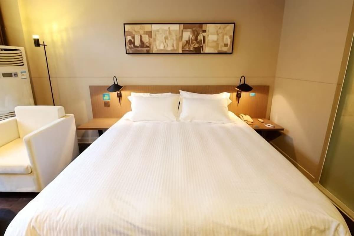 A hotel room with a bed, a chair, a nightstand, a lamp, and a wall-mounted phone.