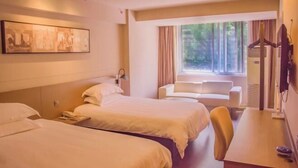 Ein Hotelzimmer mit zwei Betten, einem Schreibtisch, einem Stuhl, einem Fenster mit Vorhängen und einem an der Wand montierten Fernseher.