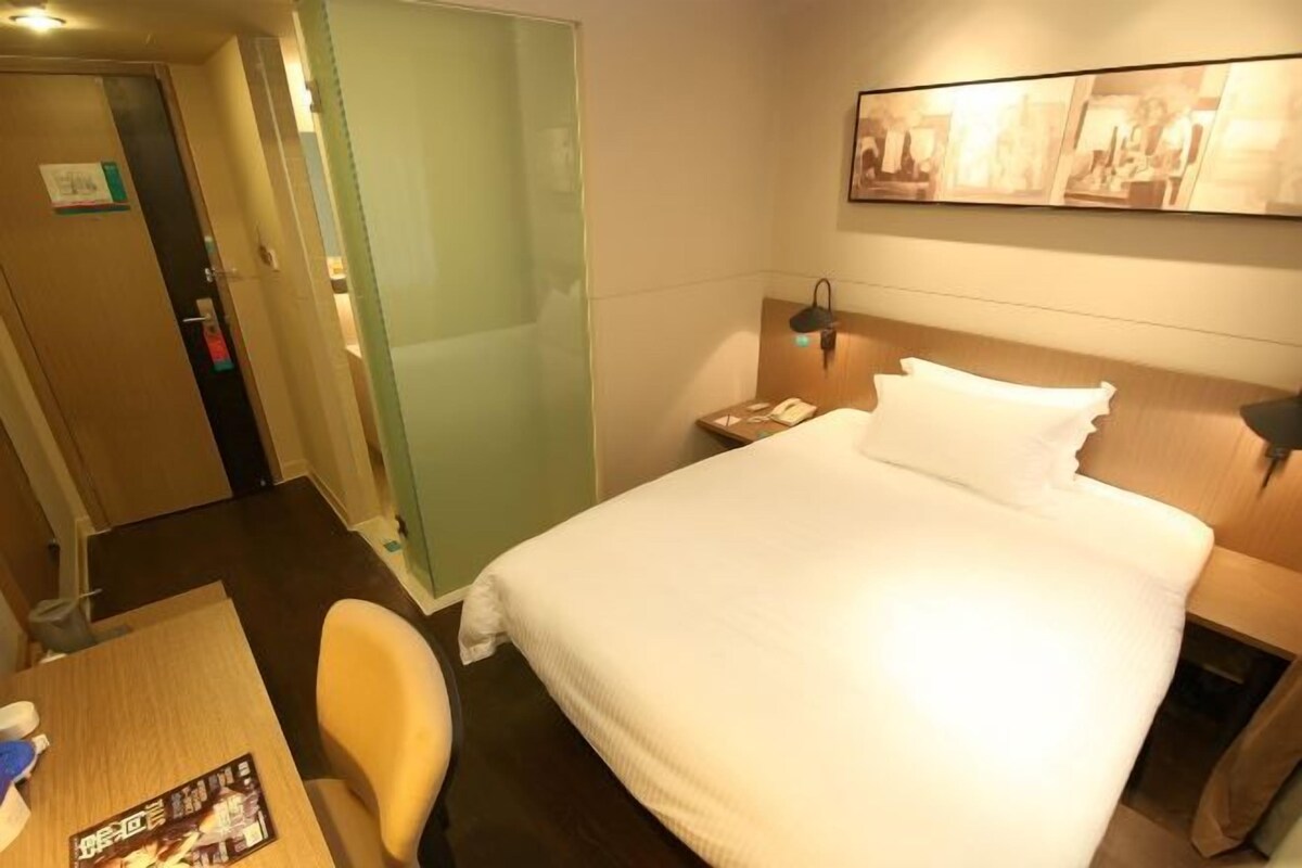 A hotel room with a bed, a desk, a chair, and a nightstand with a lamp.