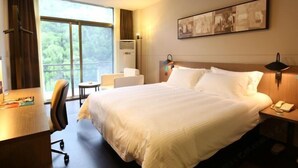Habitación de hotel con una cama grande, un escritorio, una silla, una mesita de noche con lámpara y una ventana con vistas a la vegetación.