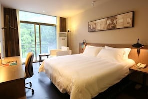 A hotel room with a large bed, a desk, a chair, a bedside table with a lamp, and a window with a view of greenery. - Jinjiang Inn Select (Lushan City)