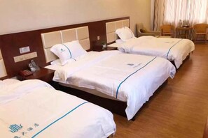 A hotel room with three beds, a wooden headboard, a small table with a lamp, and a chair. - Huating Hotel (Mojiang)