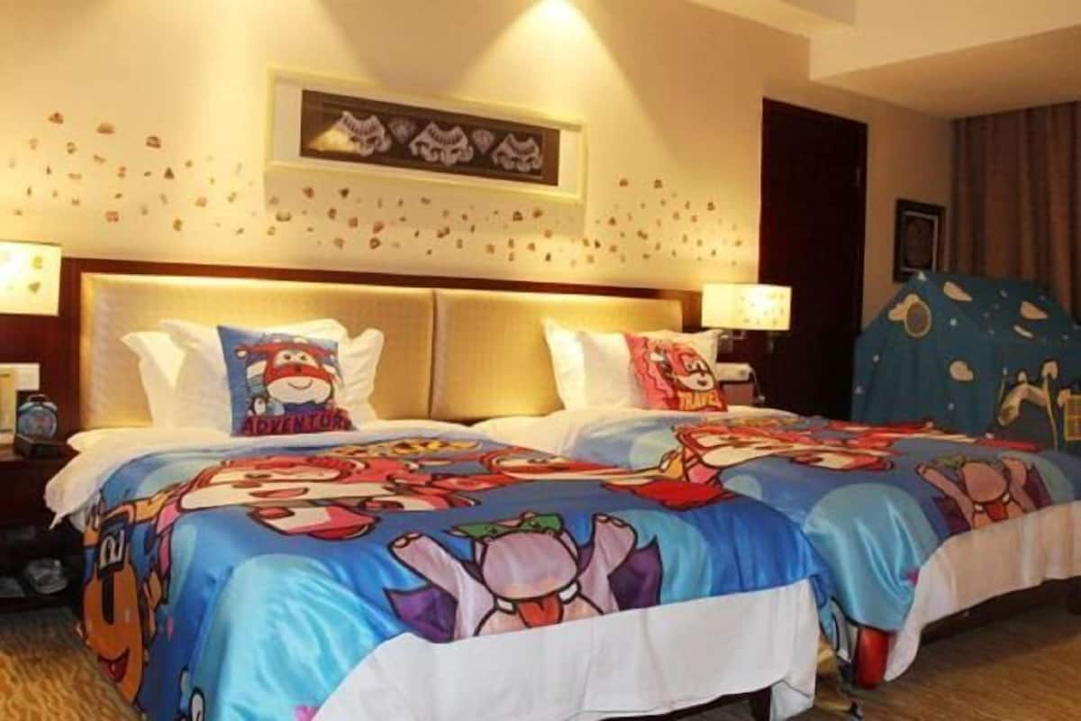 Quarto de hotel com duas camas, cada uma com colcha colorida com personagens de desenho animado. Há duas mesas de cabeceira com abajures, um relógio de parede e um quadro emoldurado sobre as camas.
