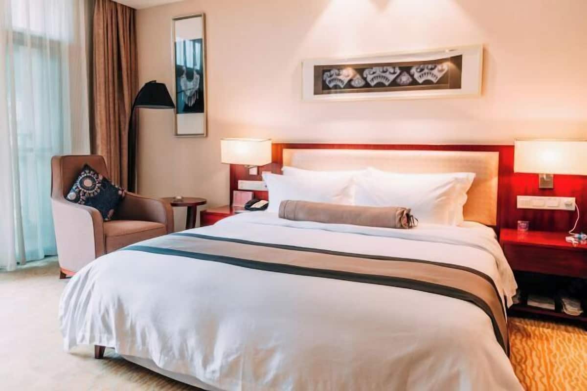 Quarto de hotel com uma cama grande, duas mesas de cabeceira com luminárias, uma cadeira e uma obra de arte na parede.