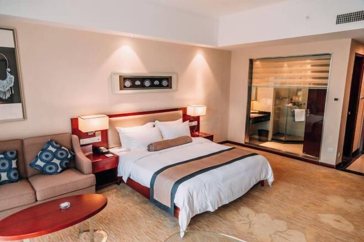 Quarto de hotel com cama, sofá, mesa de cabeceira, abajur, relógio de parede e porta corrediça para o banheiro.