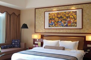Habitación de hotel con una cama grande, dos lámparas de noche, un sofá con cojines estampados, un cuadro en la pared y una ventana con cortinas.