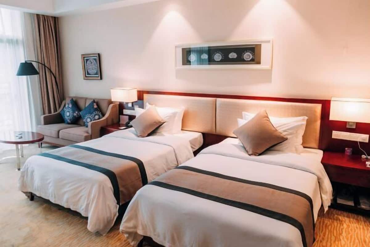 Quarto de hotel com duas camas, um sofá, uma mesinha e uma luminária.