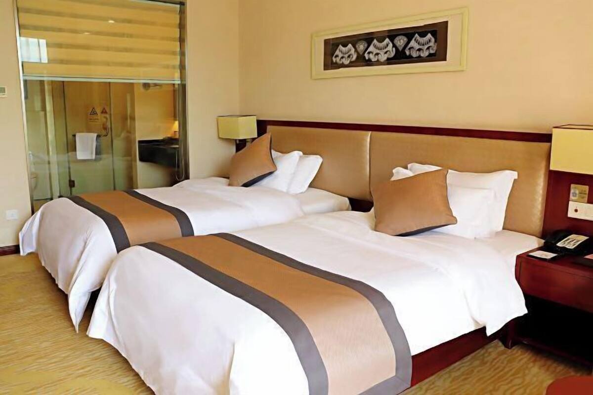 Quarto de hotel com duas camas, uma porta de vidro, um quadro na parede e uma mesa de cabeceira com abajur.