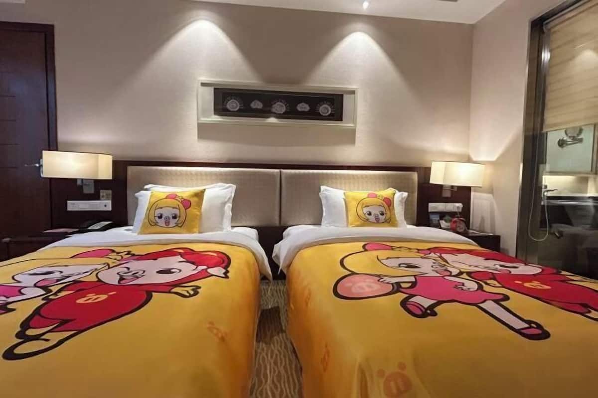 Quarto de hotel com duas camas, cada uma com edredom estampado com personagens de desenho animado, cabeceiras, abajures e um relógio de parede.