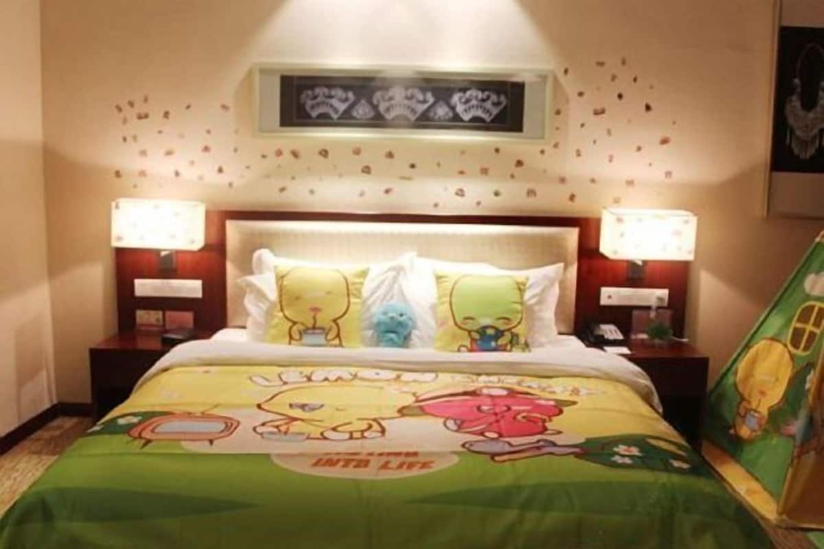 Quarto de hotel com cama decorada com estampas de personagens de desenhos animados, duas luminárias de cabeceira, uma mesa de cabeceira e um adorno na parede.