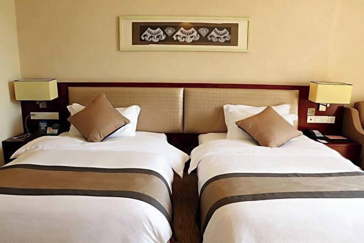 Quarto de hotel com duas camas, cada uma com cabeceira, mesas de cabeceira, luminárias e uma obra de arte emoldurada na parede.