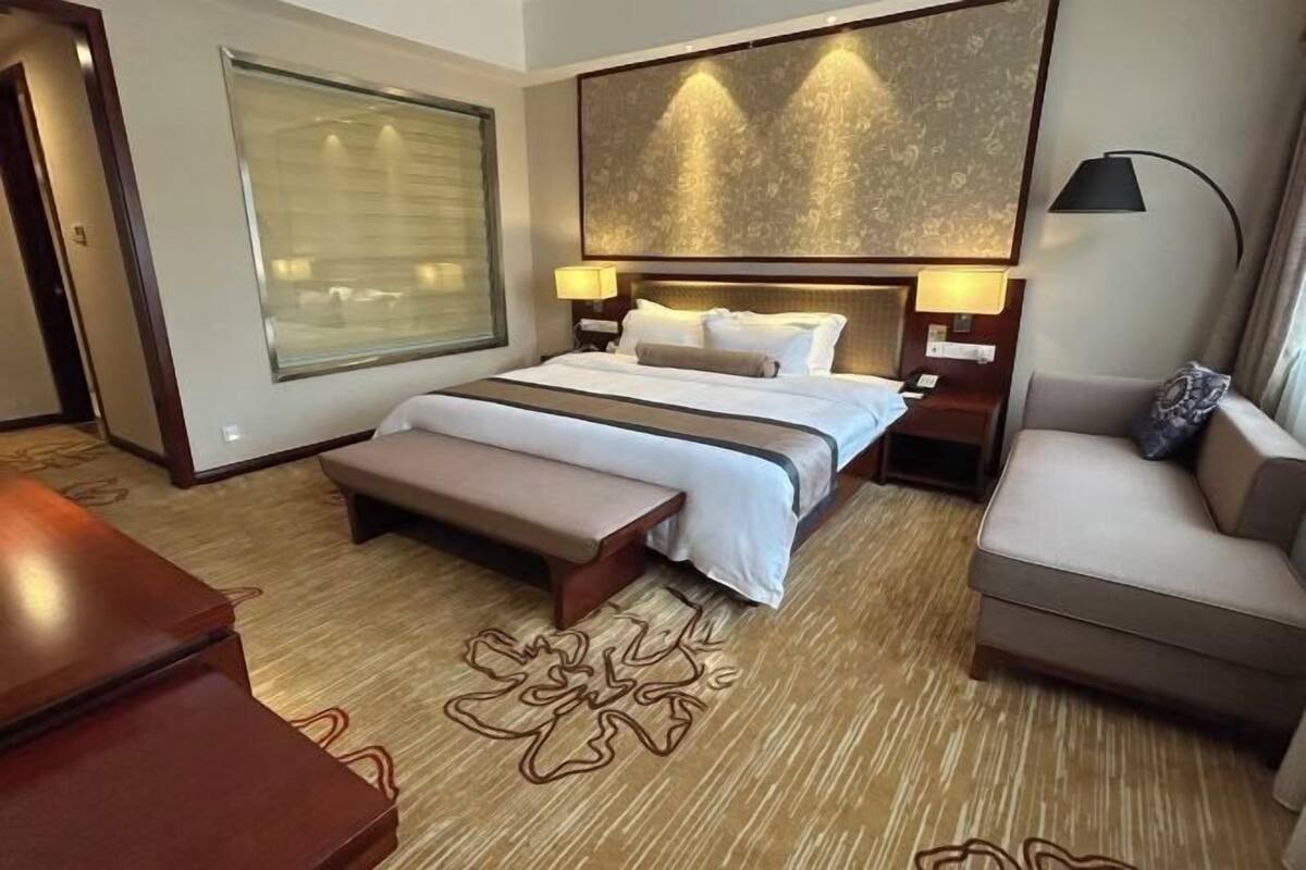 Quarto de hotel com uma cama grande, um banco, um sofá, uma escrivaninha e uma janela ampla com persianas.