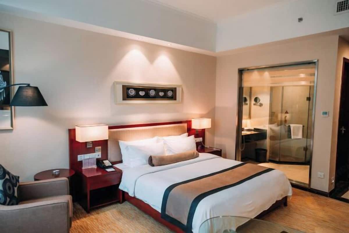 Quarto de hotel com uma cama, mesas de cabeceira, uma luminária, um quadro na parede e uma porta deslizante para o banheiro.