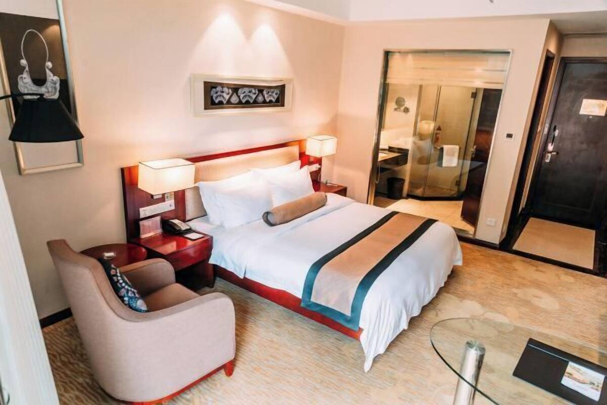 Quarto de hotel com cama, mesas de cabeceira, uma cadeira, uma mesa de vidro e um guarda-roupa.