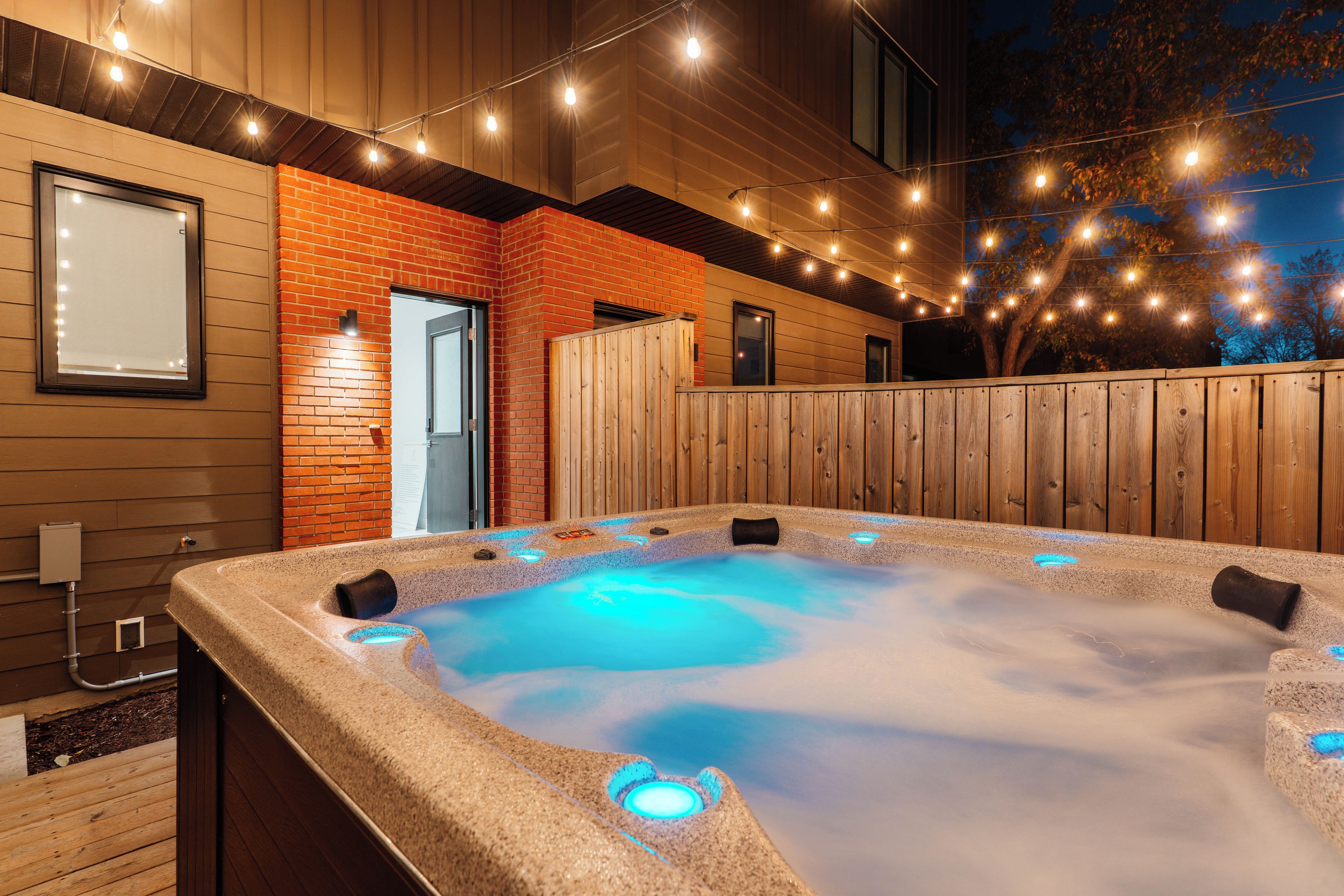 Un bain à remous avec une terrasse en bois, entouré d’une clôture en bois et illuminé de guirlandes lumineuses.