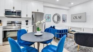A modern kitchen with white cabinets, stainless steel appliances, and a dining area with blue chairs and a round table. - The Olaf in Trendy Killarney - Close to Everything (Calgary)