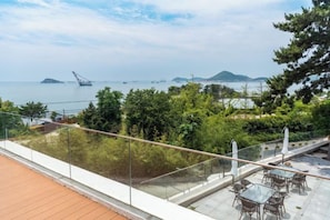 A terrace with a glass railing overlooking a body of water, greenery, and distant islands.