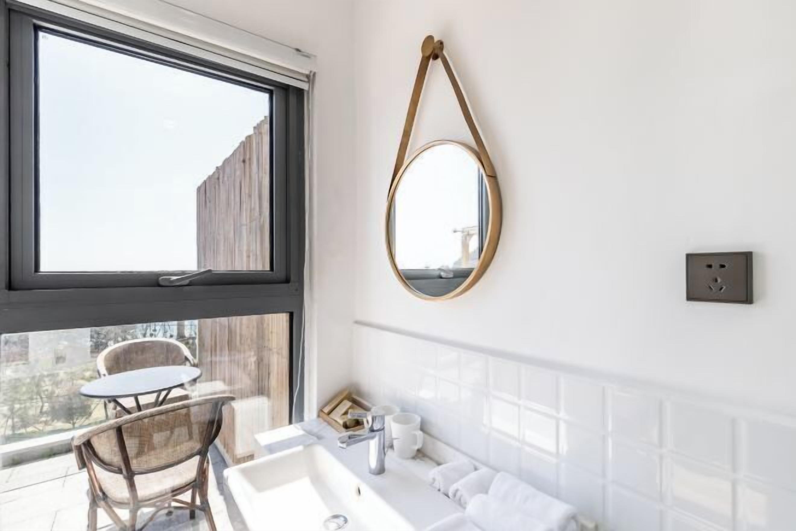 A modern bathroom with a large window, a round mirror, a white sink, and a chair by the window.