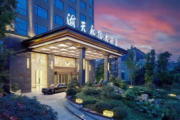 A hotel entrance with a covered walkway, a car parked in front, and a well-maintained garden.