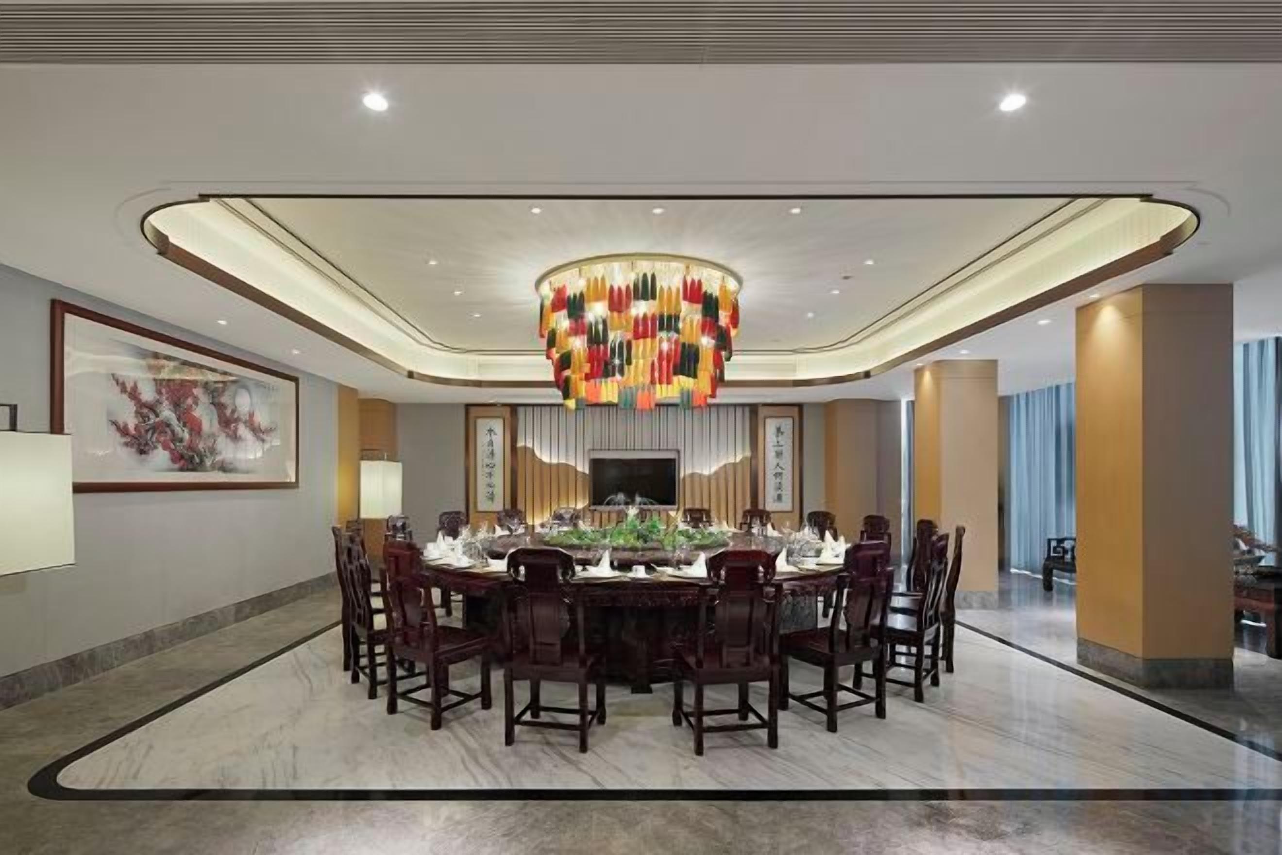 A spacious dining area with a round table, multiple chairs, a decorative chandelier, and a television screen.