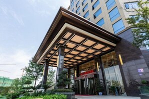 Entrada moderna de hotel com área coberta, portas de vidro e jardim ajardinado.