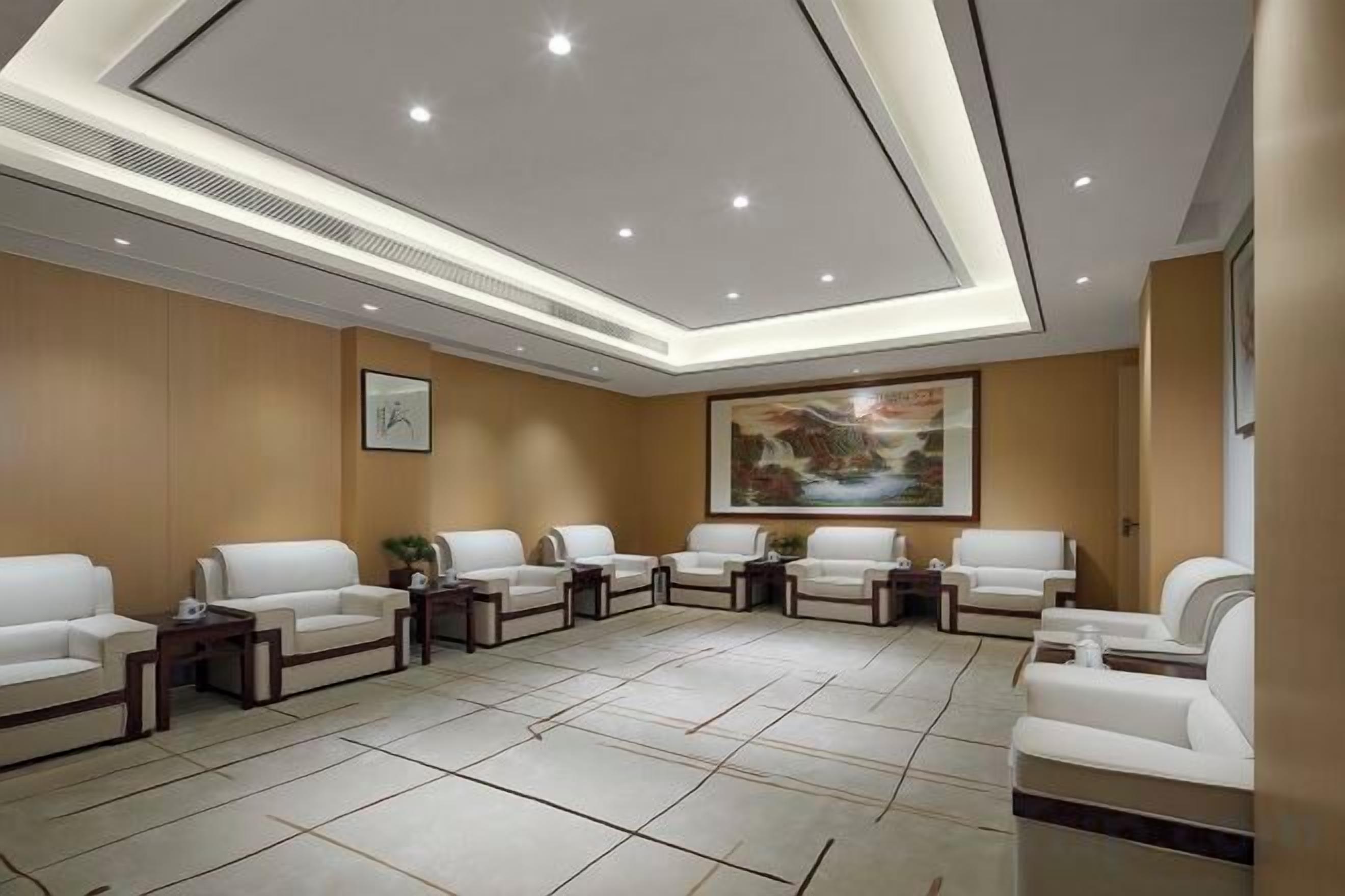 A spacious waiting area with multiple white sofas arranged in a semi-circle, a large painting on the wall, and a recessed ceiling with integrated lighting.