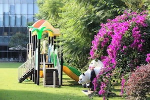Un parque infantil con un tobogán y estructuras de escalada, rodeado de abundante follaje y plantas con flores.