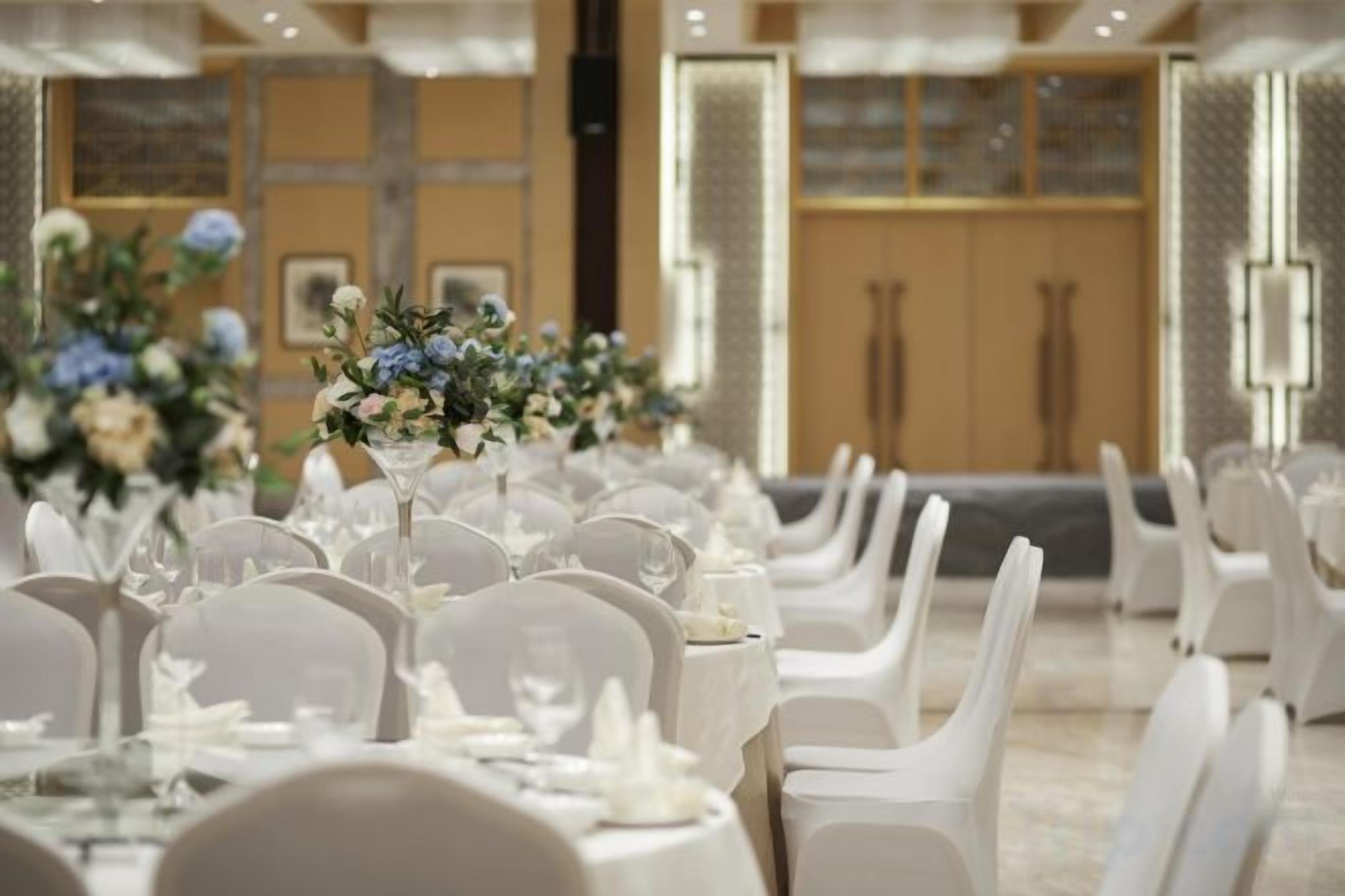Een balzaal met ronde tafels gedekt voor een formeel evenement, met witte stoelen en bloemstukken als decoratie.