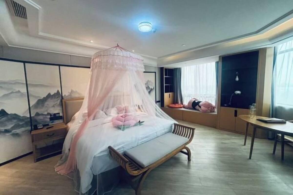 Een slaapkamer met een hemelbed, een houten bankje, een nachtkastje, een spiegel en kunst aan de muur.