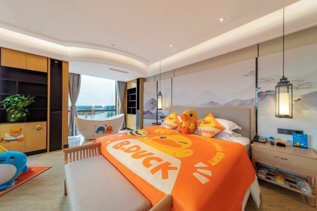 Une chambre d’hôtel avec un lit agrémenté d’une couvre-lit à motifs de canards, une table de chevet avec une lampe, une vue sur la mer et une grande fenêtre.