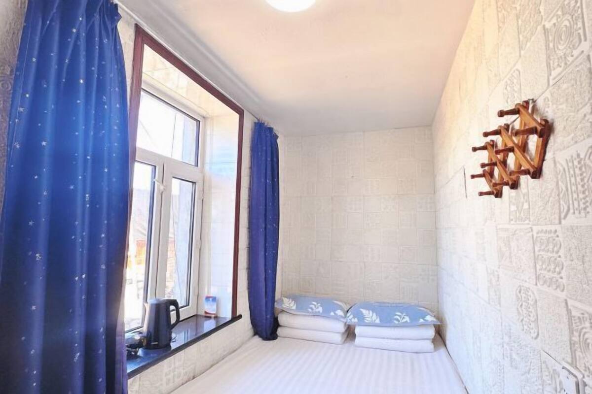 A small room with a bed, a window with curtains, a wall-mounted light fixture, and a small table with a kettle.