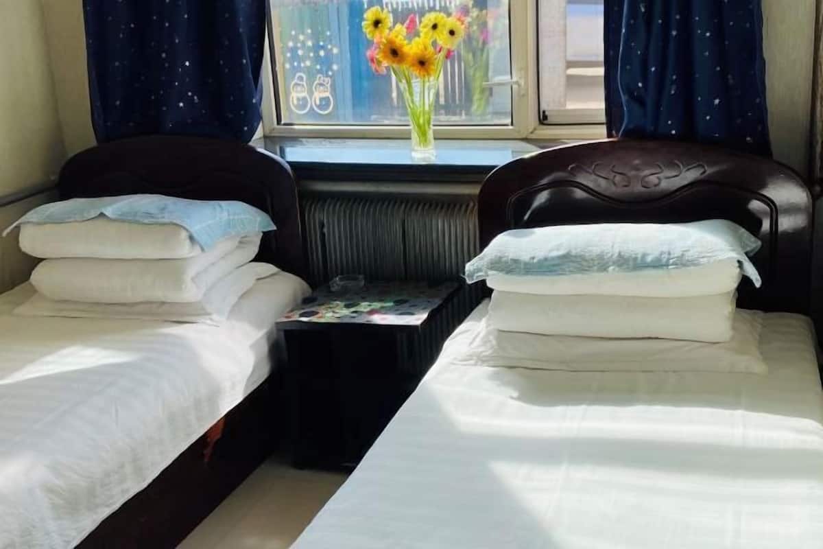 A room with two single beds, each with a pillow and a blanket. There is a small table with a floral arrangement and a window with curtains.