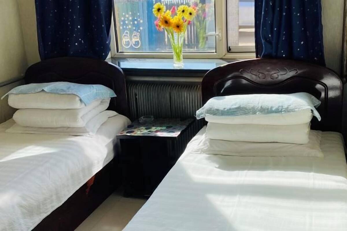 A room with two single beds, each with a pillow and a blanket. There is a small table with a vase of flowers between the beds.