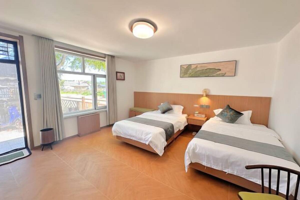 A hotel room with two beds, a window with curtains, a bedside table with a lamp, and a painting on the wall.
