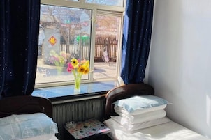 Una habitación con cama, una mesita con un jarrón de flores y una ventana con cortinas.