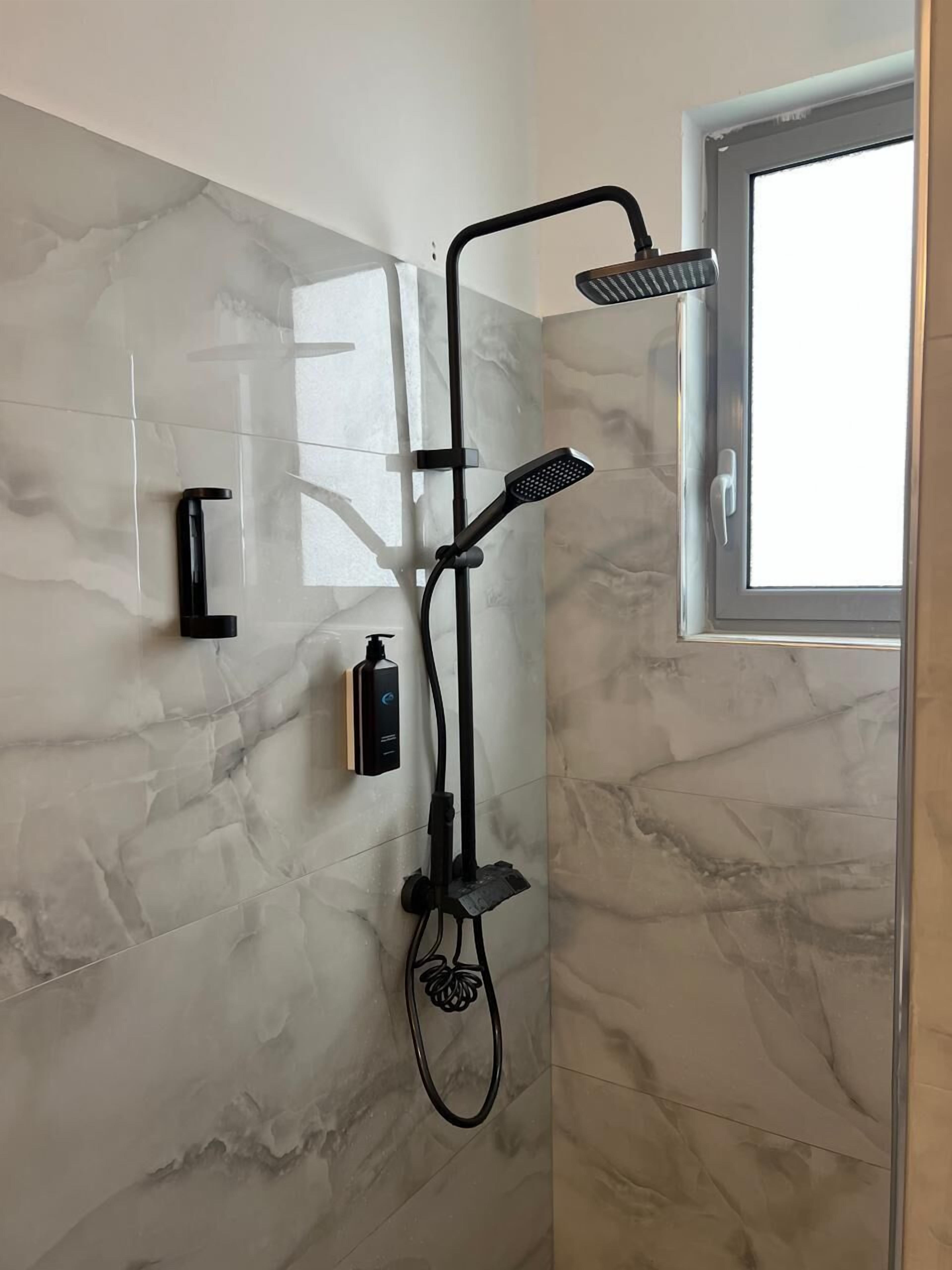 Un baño moderno con ducha, cabezal de ducha de mano, dispensador de jabón de pared y ventana para luz natural.