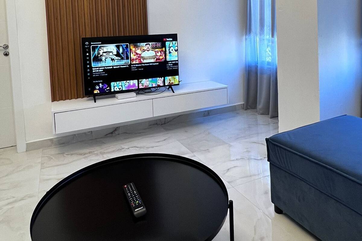 A modern living room with a flat-screen TV mounted on a white wall, a black round table with a remote control, a blue sofa, and a window with curtains.