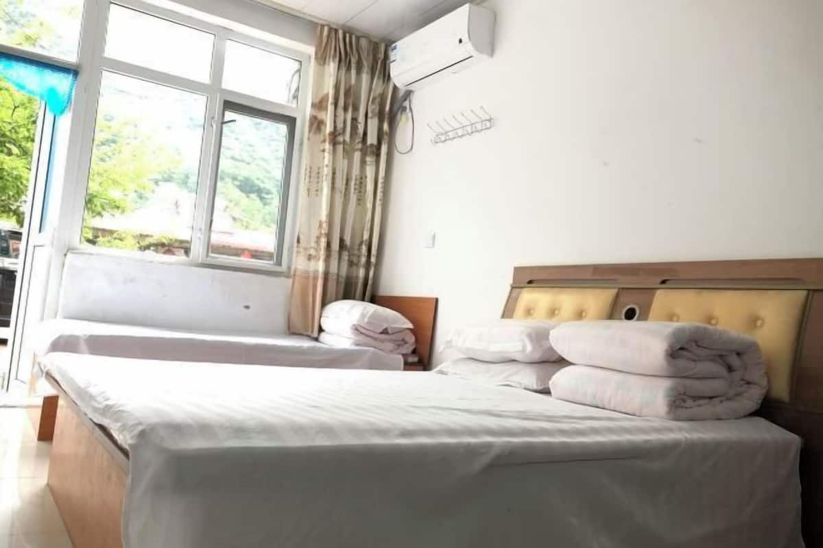 A hotel room with two beds, a window with curtains, an air conditioning unit, and a bedside table.