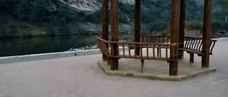 A wooden gazebo with benches, located near a body of water with a mountainous backdrop.