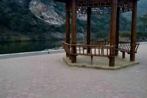 A wooden gazebo with benches, located near a body of water with a mountainous backdrop.